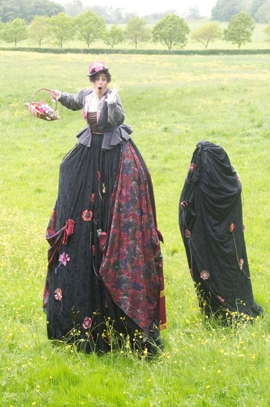 The Victorian Flower Seller character on stilts in a field holding a basket of flowers